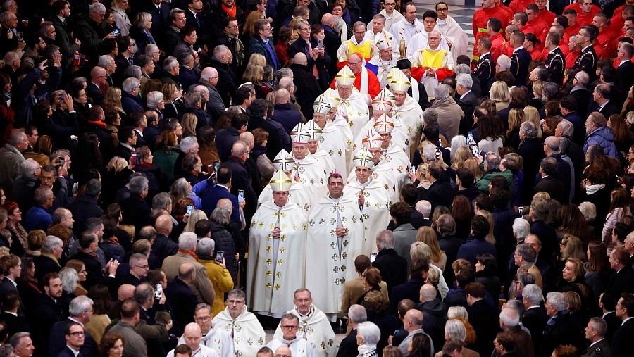 Procesión de sacerdotes en Notre Dame, observada por una multitud que usa sus móviles para grabar. Un grupo en uniformes rojos participa en el evento.