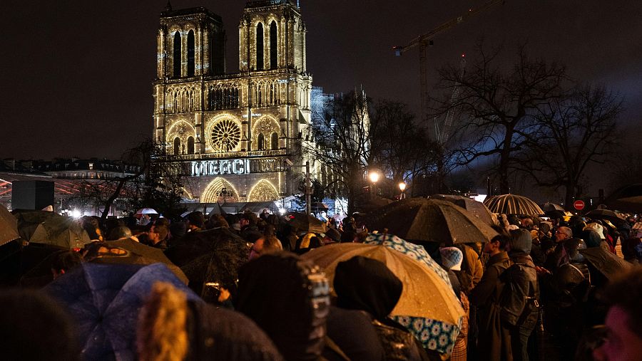 Multitud bajo paraguas observa la Catedral de Notre Dame iluminada, donde se proyecta la palabra 
