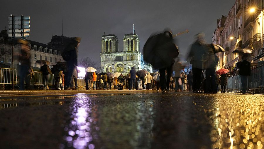 Impresionante vista nocturna de Notre Dame, reflejada en el suelo mojado. Multitud con paraguas ante la catedral iluminada.