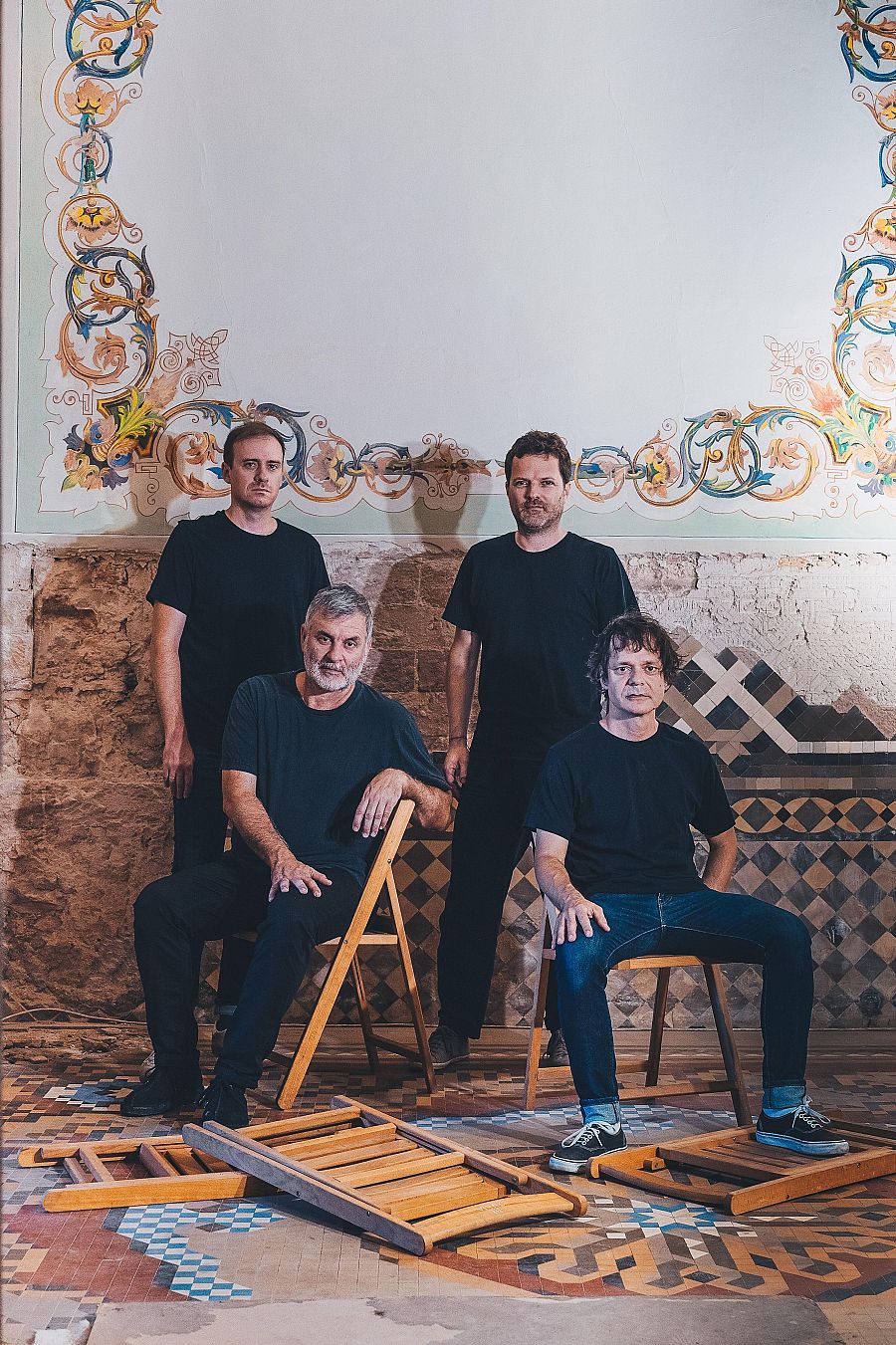 Fotografía de cuatro hombres con camisetas negras en un espacio interior con paredes de piedra, suelo embaldosado y sillas plegables, algunas rotas.