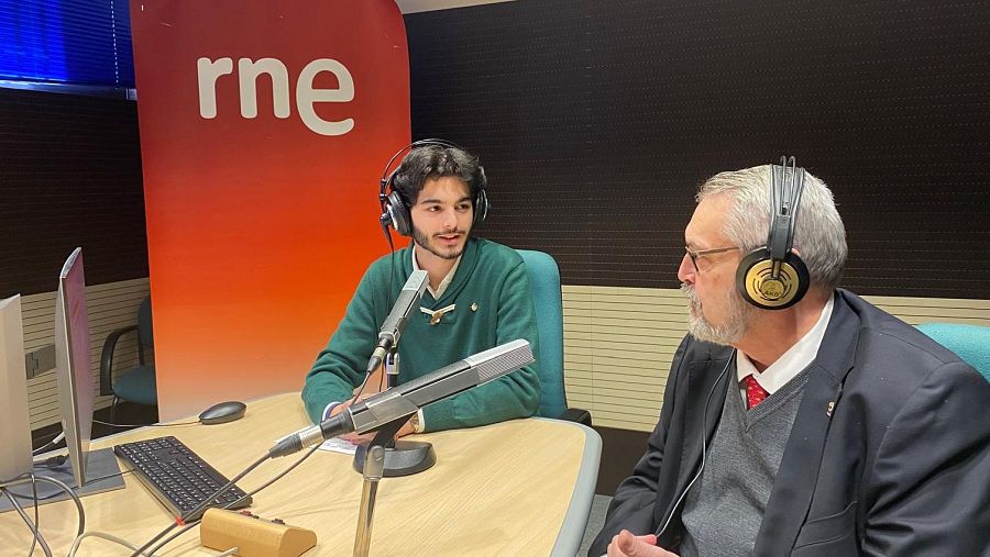En un estudio de RNE Málaga, dos hombres, uno joven con jersey verde y otro mayor con traje oscuro, participan en un programa de radio. Se aprecian micrófonos y auriculares.