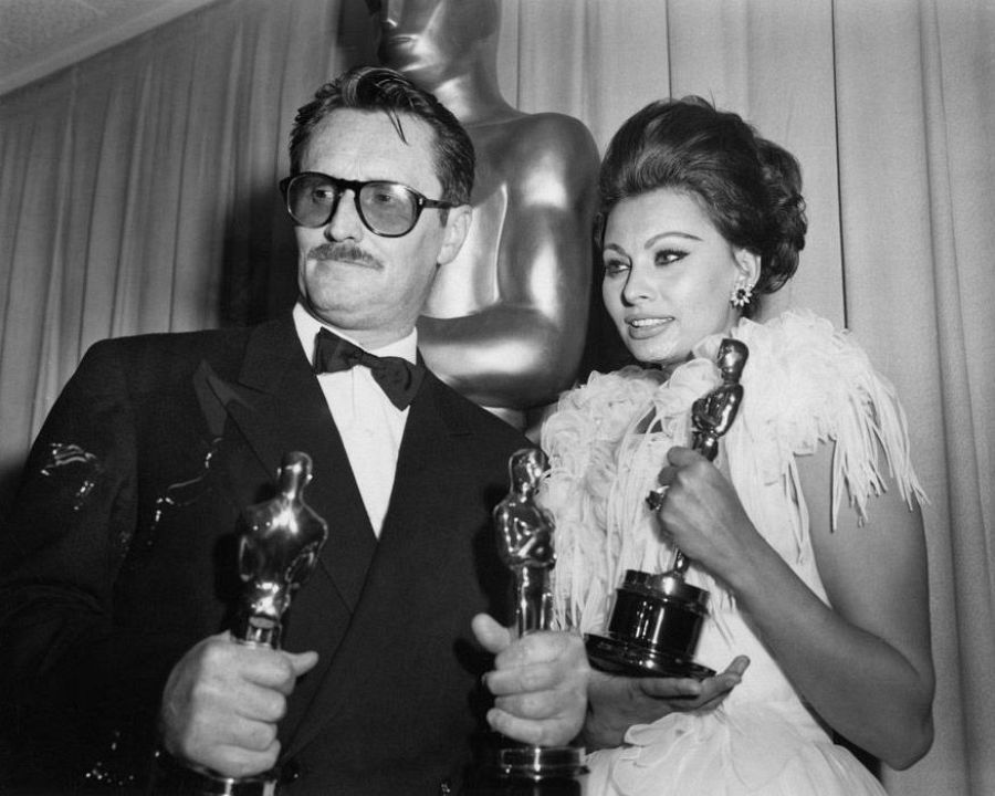 Pietro Germi y Sophia Loren, en blanco y negro, durante los Oscar de 1962.  Ambos sostienen dos estatuillas cada uno, vestidos de gala.