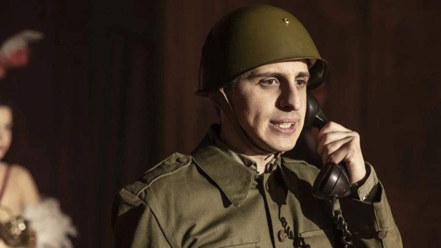 Óscar Lastre, en uniforme militar, interpreta a Miguel Gila usando un teléfono antiguo en una escena teatral.  Al fondo, una mujer con vestido brillante.