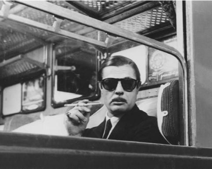 Marcello Mastroianni, con gafas de sol y traje, fuma un cigarrillo mientras mira a la cámara con expresión seria desde el interior de un vehículo.