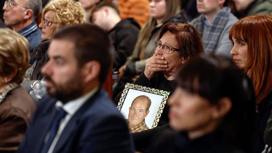 Mujer con gafas sostiene con ambas manos un retrato de un hombre mayor, mostrando profunda tristeza en un acto conmemorativo.