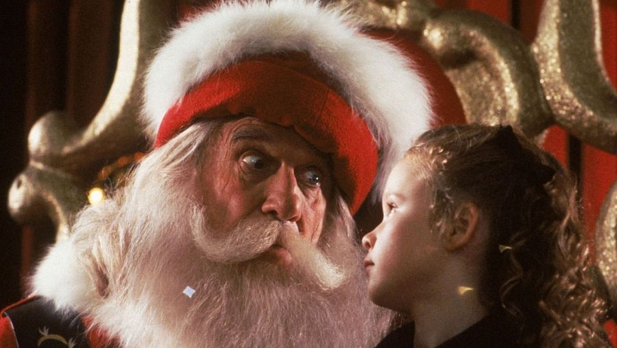 Leslie Nielsen, caracterizado como Santa Claus, comparte una escena con una niña en un decorado navideño.  La expresión del actor es cómica.