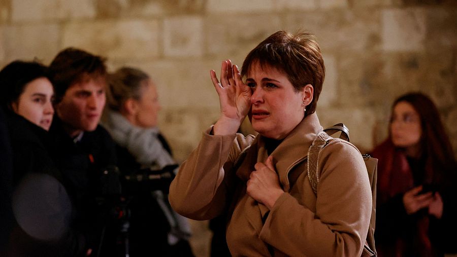 Mujer con abrigo beige y mochila, visiblemente afectada, sale de una catedral.  Secándose las lágrimas, refleja el dolor por una pérdida.