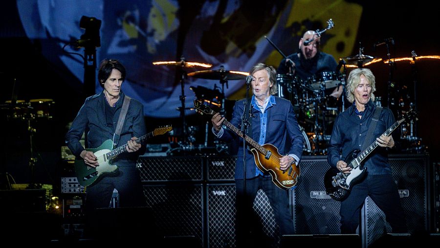 Paul McCartney en concierto: tocando el bajo y cantando, acompañado por dos guitarristas y un batería. Iluminación tenue, escenario con amplificadores.