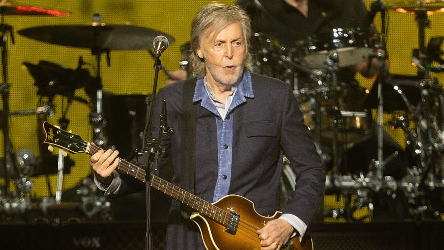Paul McCartney toca un bajo marrón brillante en un escenario con fondo amarillo. Lleva camisa azul y chaqueta oscura, y canta mientras toca.