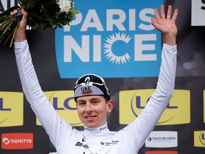 Ciclista con maillot blanco celebra victoria, sosteniendo flores y saludando. Se ve un panel con el logo de la carrera 