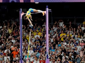 Atleta en pleno salto con pértiga, cuerpo arqueado, uniforme azul y amarillo, multitud en las gradas.