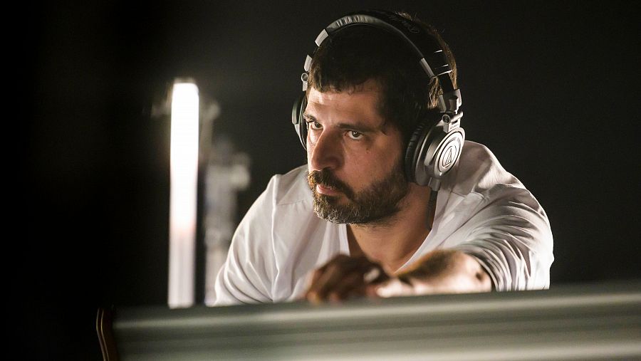 Marcel Bagés, amb auriculars, es concentra en una tasca en una taula de mescles.  La seva expressió seriosa i el fons fosc ressalten el seu enfocament a la producció d'àudio. Porta una camisa blanca i barba.