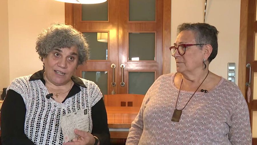 Dos mujeres maduras conversan en un interior; una sostiene un documento, la otra lleva un micrófono.  Se observa una puerta de madera con cristal.