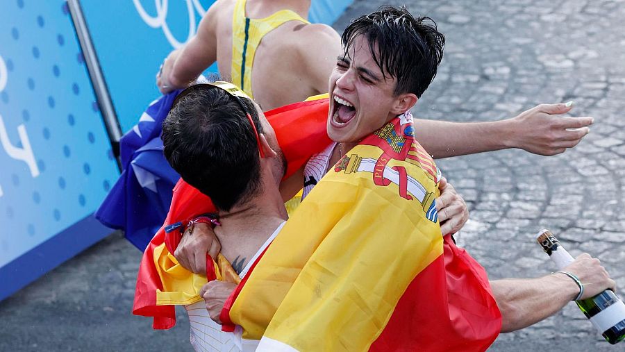 Emotiva celebración de atletas tras ganar el oro olímpico en marcha atlética. Un atleta con la bandera de España, y otro abrazándolo con una botella, festejan la victoria ante un panel con los anillos olímpicos.