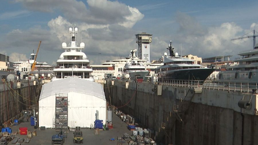 Dique seco con tres yates, uno cubierto con lona blanca, y equipo de mantenimiento visible. Instalaciones de reparación naval.