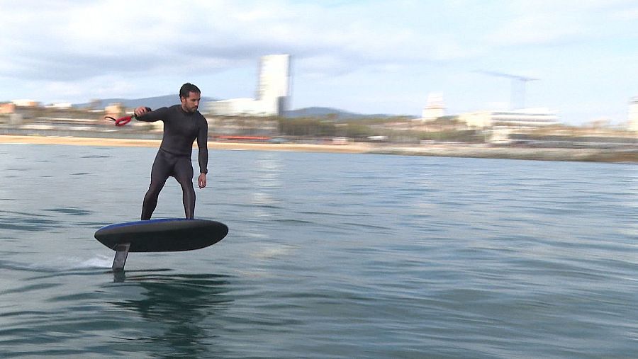 Hombre en traje de neopreno practica flyboard, elevándose sobre el agua con un dispositivo en la mano; al fondo, línea costera con edificios.