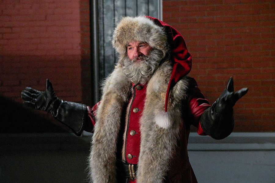 Santa Claus, con traje rojo y ribetes de piel, guantes negros y gesto de bienvenida, sobre fondo de ladrillo rojo.