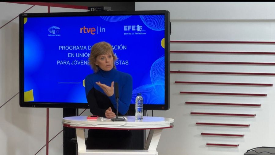 Presentadora de RTVE hablando tras un atril con micrófono, botella de agua y móvil.  Fondo con pantalla mostrando logo EFE y detalles de un programa de formación para jóvenes periodistas.