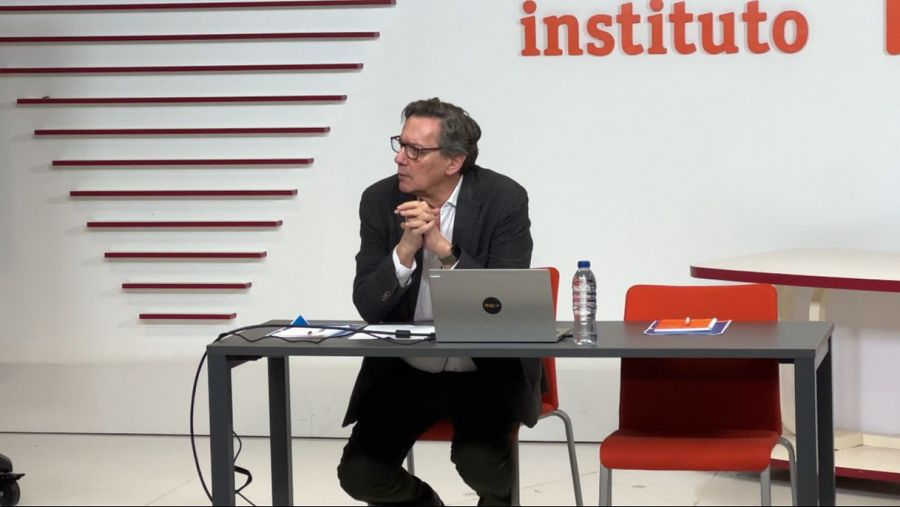 Persona con gafas y chaqueta oscura sentada a una mesa con un portátil, agua y un bloc de notas en un instituto.  Ambiente de conferencia o evento.