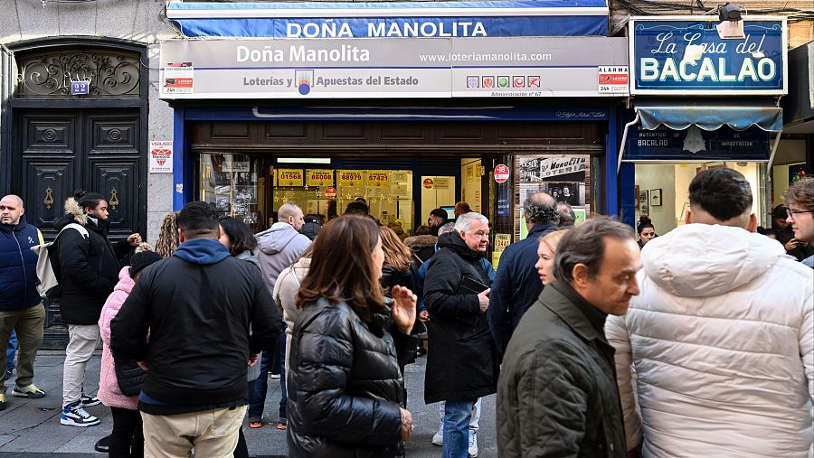 Cola de personas frente a la administración de loterías Doña Manolita en Madrid.  Se observa el letrero con el nombre y la dirección web.