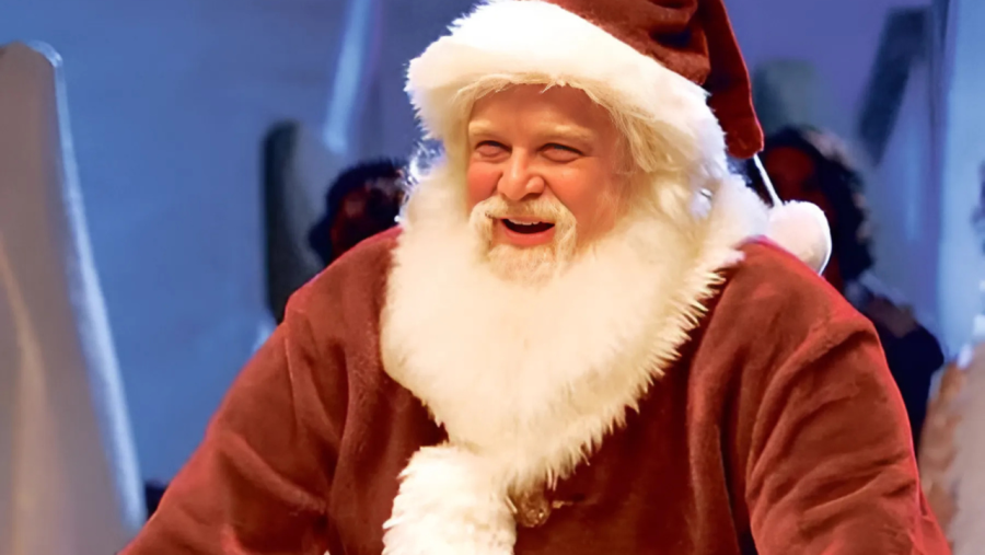 John Goodman, vestido como Santa Claus con traje rojo y blanco, sonríe ampliamente en una imagen con fondo borroso.