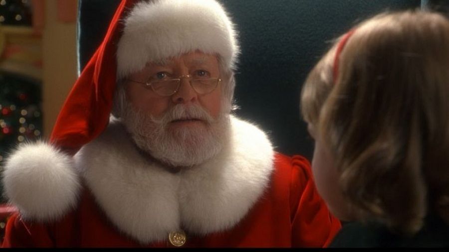 Santa Claus, interpretado por Richard Attenborough, interactúa con una niña en una escena navideña. El actor viste el traje tradicional y se encuentra sentado en un sillón.