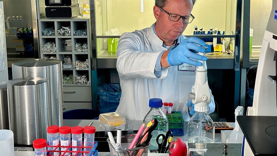 Inmunólogo en laboratorio con bata blanca, gafas y guantes azules, manipulando un instrumento de pipeteo y rodeado de material científico.