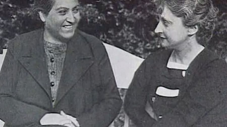 Dos mujeres, una sonriente y otra con expresión seria, sentadas en un jardín.  Imagen en blanco y negro, posiblemente de una entrevista.