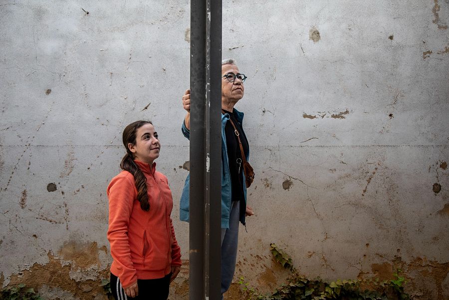 Dos mujeres, una joven y una mayor, miran hacia arriba junto a un poste metálico en un entorno exterior desgastado.  Ambas visten ropa informal.