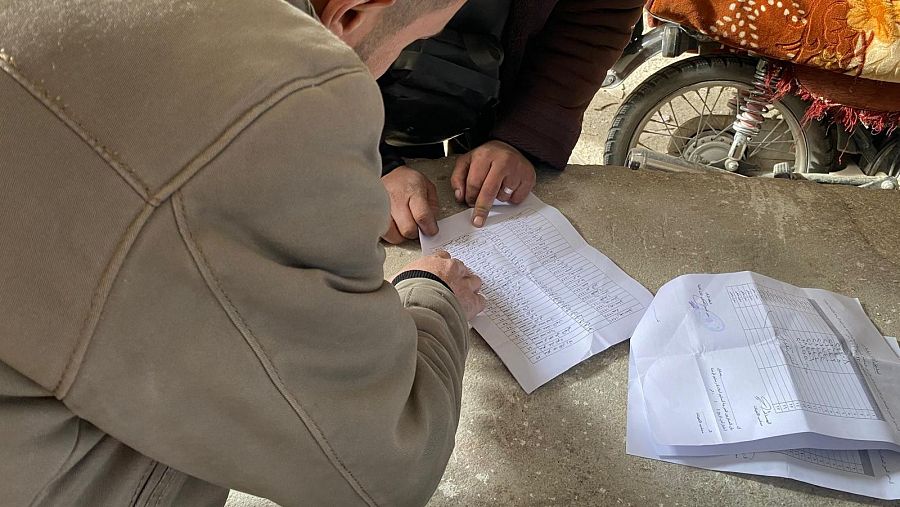 Dos personas revisan documentos, posiblemente registros oficiales, en una mesa improvisada.  Una lleva una chaqueta beige y la otra una prenda granate. Se ve parte de una motocicleta al fondo.