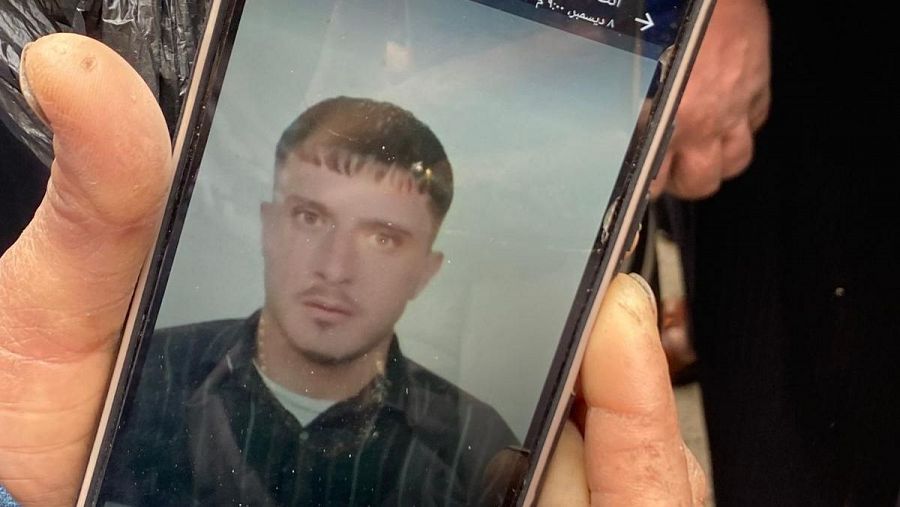 Retrato tipo carnet de un hombre con pelo corto y oscuro y camisa oscura a rayas, mostrado en un teléfono móvil. Se observa la imagen a otra persona.