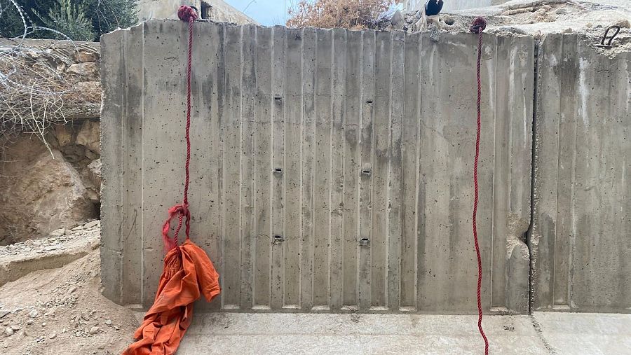 Bloque de hormigón gris con textura vertical acanalada y grieta central; dos cuerdas rojas sujetan una prenda naranja.