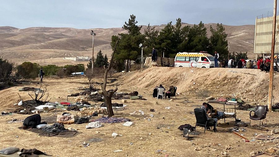 Familias refugiadas en un terreno desolado reciben asistencia médica y humanitaria.  Se observan mantas, colchones y escombros.