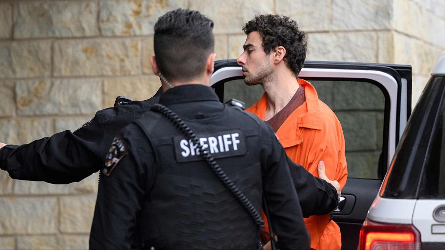 Un preso con mono naranja es escoltado por un sheriff hacia un vehículo policial frente a una pared de piedra.  Imagen que sugiere traslado a un juzgado.