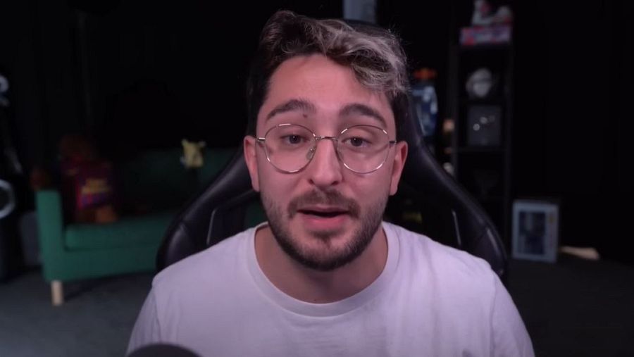 Youtuber con gafas y camiseta blanca, sentado en silla gamer, durante una transmisión en directo. Fondo oscuro con objetos borrosos.