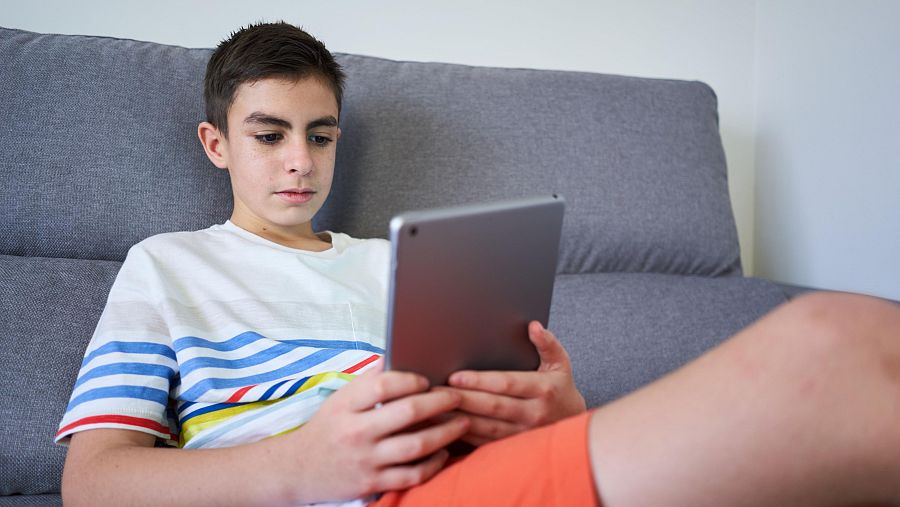 Joven concentrado mirando una tablet que sostiene con ambas manos; viste camiseta a rayas y pantalones cortos.