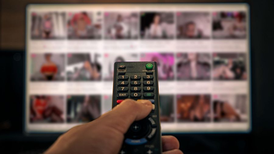 Mano sujetando control remoto frente a pantalla con miniaturas borrosas de videos con contenido para adultos.  Énfasis en la facilidad de acceso a este tipo de contenido.