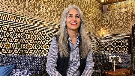 Entrevista a Fariba Ehsan, con camisa a rayas y chaleco, en un interior con decoración árabe; azulejos geométricos y florales, banco con tapizado azul y blanco.