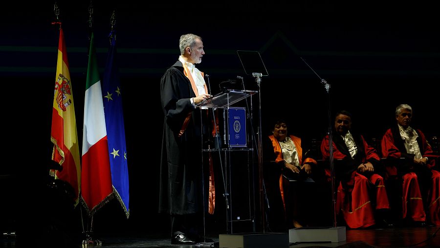 El rey Felipe VI, en toga académica, da un discurso ante banderas de España, Italia y la UE; al fondo, otras personas en togas académicas.