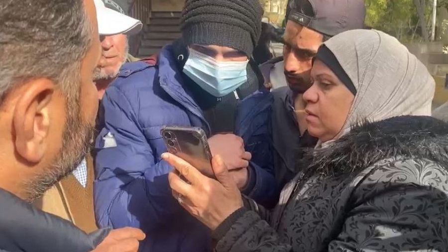 Grupo de personas observan con preocupación la pantalla de un teléfono móvil mostrado por un joven con gorro y mascarilla.  Ambiente exterior.