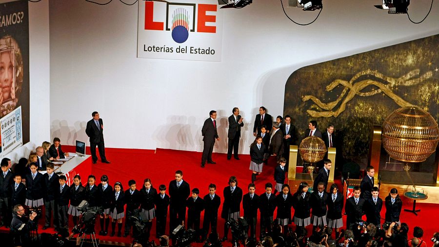 Niños uniformados en un escenario con bombos dorados, listos para el sorteo.  Se observa el logo de Loterías y Apuestas del Estado en el fondo.