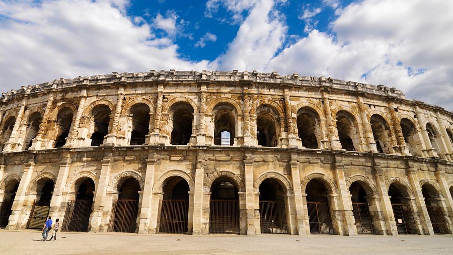 Imponente anfiteatro de piedra beige con múltiples arcos, parcialmente en ruinas en su interior pero con la parte superior bien conservada. Se aprecian dos personas en la parte inferior.