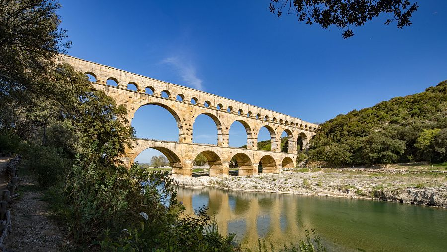 El Acueducto de Nimes en el puente de Gard: múltiples arcos de piedra sobre un río, con vegetación y un cielo despejado. Imagen de un documental.