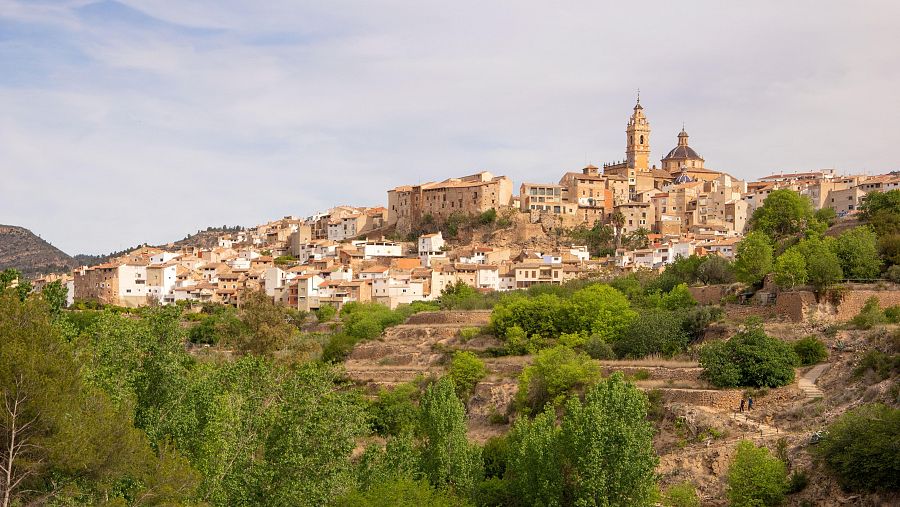 Vista panorámica de un pueblo en una ladera, con casas, iglesia, vegetación y terrazas de cultivo.  Cielo parcialmente nublado; dos personas caminan por un sendero.