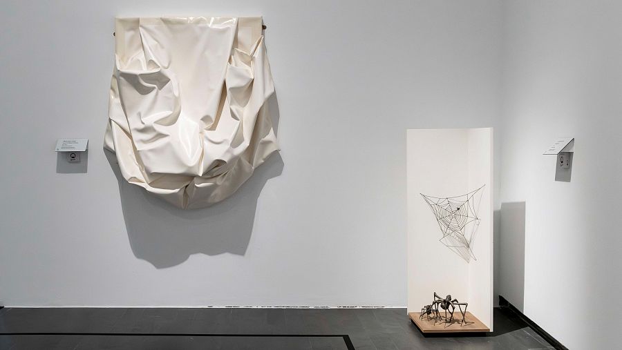 Sala de exposiciones con una pieza textil blanca arrugada, una escultura de araña y sus telarañas, ambas con etiquetas informativas. Suelo de baldosas oscuras e iluminación uniforme.