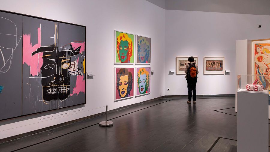 Sala de exposiciones con obras abstractas y serigrafías de Marilyn Monroe. Una persona con mochila naranja observa las obras desde la parte trasera.
