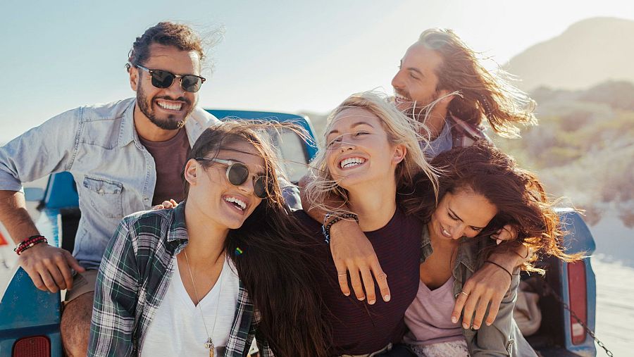 Cinco jóvenes, felices y abrazados, disfrutan de un día soleado junto a una camioneta azul en un paisaje arenoso.  Ropa informal de verano.