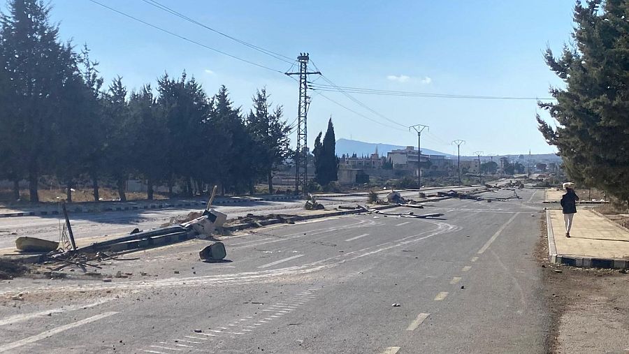 Carretera siria deteriorada con escombros, postes caídos y una torre eléctrica cercana.  Una persona camina por la acera.