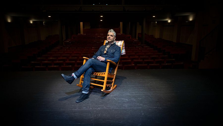 Artista sentado en mecedora de madera en teatro vacío con butacas rojas. Viste camisa vaquera y pantalones oscuros.  Imagen transmite soledad y tranquilidad.