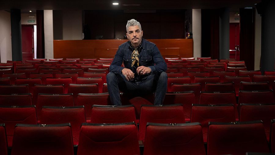 Actor sentado en un teatro vacío con butacas rojas, viste camisa vaquera, pantalones vaqueros y pañuelo.  Ambiente tranquilo.